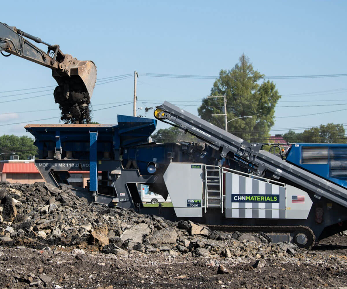 An excavator dumping broken asphalt into a CH+N Materials truck